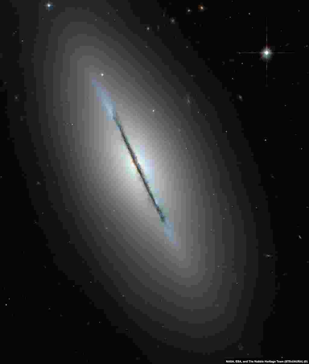 На этом снимке галактики NGC 5866 (также известной как «Веретено») можно подробно рассмотреть галактику с необычного ракурса - с ребра. Вдоль плоскости галактики четко различимы темные области космической пыли. В центре, вокруг светящегося галактического ядра можно заметить едва различимое красное сияние - это балдж, скопление плотно расположенных старых звезд.